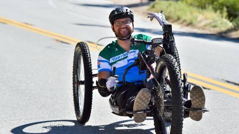 A man rides a recumbent tricycle on pavement during the Tour de Big Bear event. He waves at the camera and smiles.