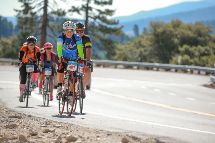 A group of cyclists ride along a roadway during the Tour de Big Bear. The cyclist at front smiles at the camera.