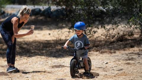 A young boy riding a Stacyc bicycle while wearing a blue helmet - his mother is standing off to the side recording him.