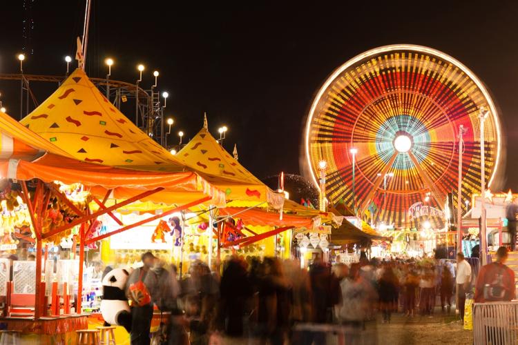 A large carnival with a ferris wheel and vendor tents - the ferris wheel is brightly lit with colors of red, blue, and yellow