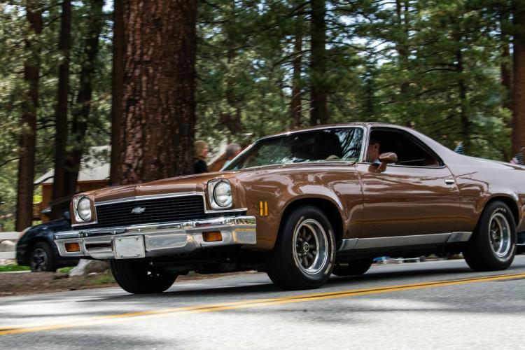 A photo of a brown antique Chevy car driving on a road - there is a dense forest of tall trees in the background.