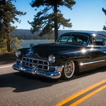An antique Hudson car driving on the road in Big Bear Lake - the mountains, trees, and lake are visible in the background.