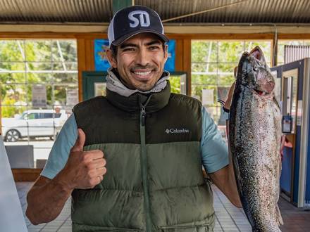 A male with a fishing jacket holding a large trout while giving a thumbs up and smiling during the Fishin' For $50K event.