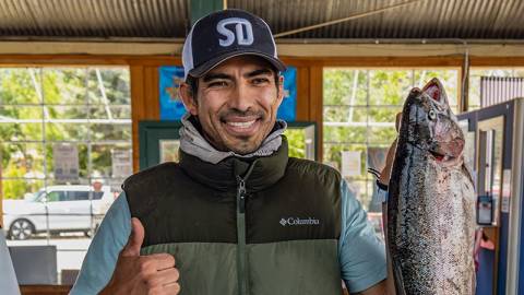 A male with a fishing jacket holding a large trout while giving a thumbs up and smiling during the Fishin' For $50K event.