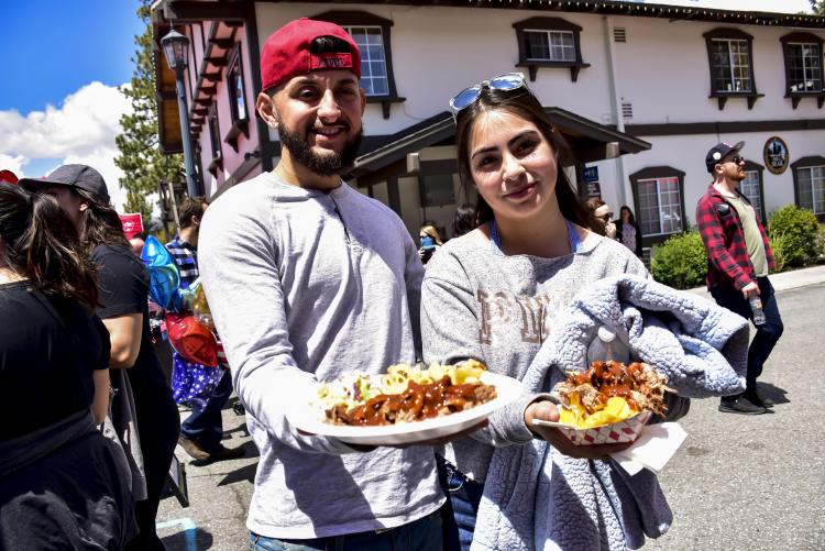 A man and woman stand in the street at the Grill & Chill BBQ Festival in Big Bear Lake, CA hold two plates of food forward.