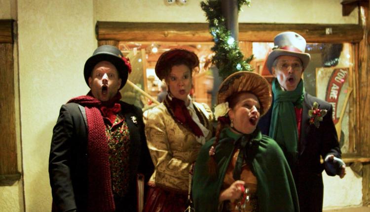A photo of small group of four people, they are Christmas Caroling, their outfits are colored with gold, red, and green.