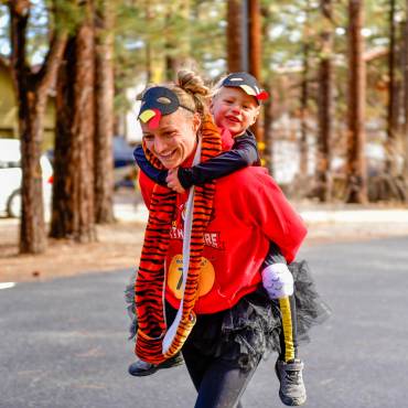 A photo of a mother with a turkey mask racing while carrying her child on her back during the Thanksgiving Turkey Trot event