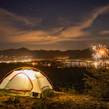 An illuminated camping tent on a hill overlooks Big Bear and the 4th of July fireworks show