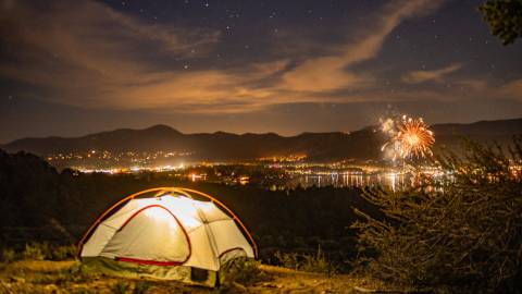 An illuminated camping tent on a hill overlooks Big Bear and the 4th of July fireworks show