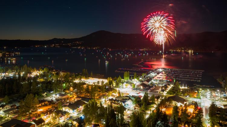 A red firework explodes above Big Bear Lake on the 4th of July. Boat and city lights are visible in this night scene.