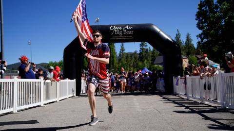 A runner carries an American flag during the Big Bear Lake 4th of July Fun Run. It's a blue bird day and fans cheer him on.