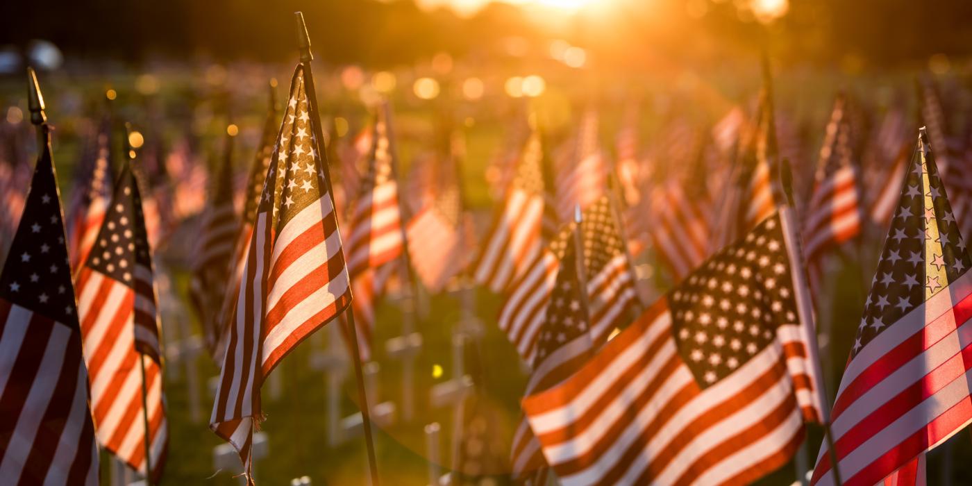 A field of white crosses topped with American Flags honoring fallen soldiers, the sun gently glaring onto the flags.