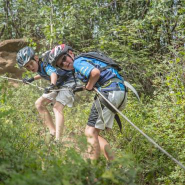Two kids wearing safety gear descending a hillside using a climbing rope - the lush green forest is surrounding them.