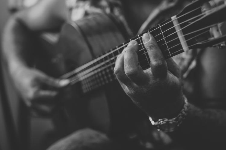 A black and white photo of someone holding and playing an acoustic guitar, the hand further away is holding a guitar pick.