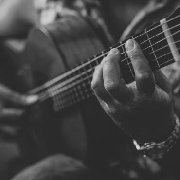 A black and white photo of someone holding and playing an acoustic guitar, the hand further away is holding a guitar pick.