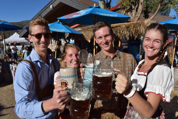 A group of four posing for a photo while holding beer mugs, they are in dressed in traditional Oktoberfest clothes.