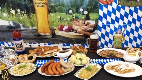 Platters of German food on a table including kielbasa, sauerkraut, soft pretzels, buns, and steins of beer