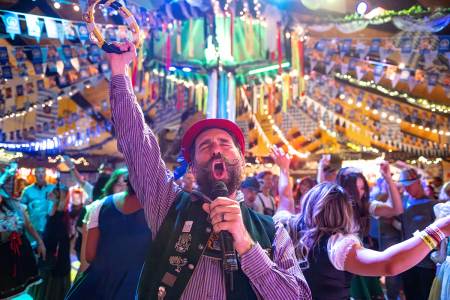 A male singer dressed in traditional German Oktoberfest attire seen singing into a microphone - a large crowd is behind him.