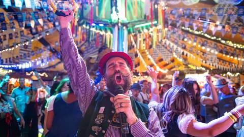 A male singer dressed in traditional German Oktoberfest attire seen singing into a microphone - a large crowd is behind him.