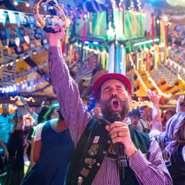 A male singer dressed in traditional German Oktoberfest attire seen singing into a microphone - a large crowd is behind him.
