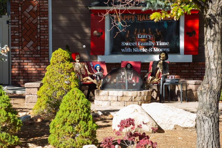 A 2025 Scarecrow & Dia de Los Muertos Display - there are skeletons relaxing on red patio chairs next to a fire pit.