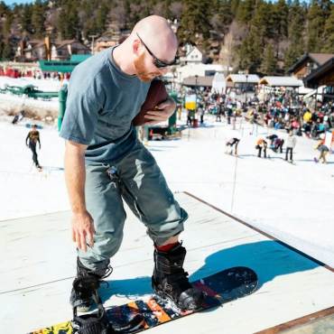 A photo of a male snowboarder holding a football - there is a crowd in the distance at the base of Bear Mountain Ski Resort.