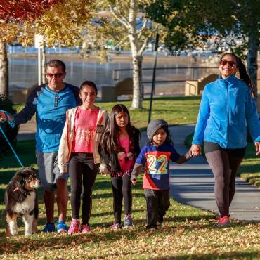 A family of five with three small children and a dog walking at the park walking through fallen leaves during Autumn.