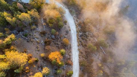 A photo of clouds slightly covering the hills below, a path is visible underneath as well as fall colored leaves on trees.