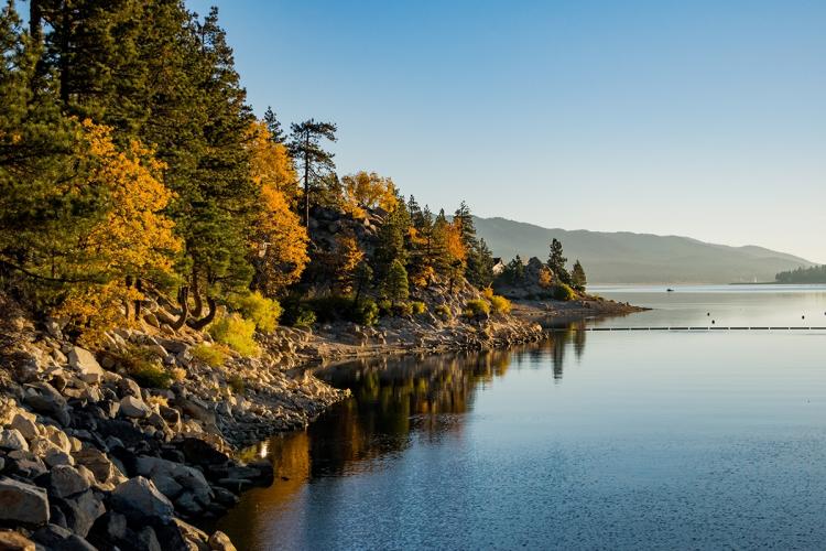 A photo taken along the Big Bear Lake waterfront, the sunset gently illuminating the fall colored trees and the beach side.