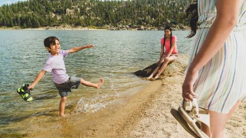 A boy splashes the water while playing on the shoreline of Big Bear Lake. His older sister and mother look on.