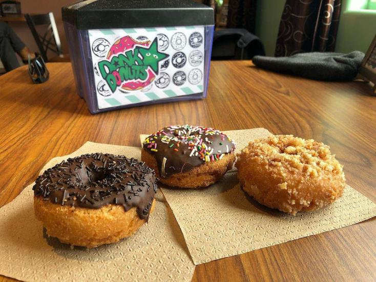 Three donuts sit on a napkin on a table. A napkin holder with the Dank Donuts logo sits in the background.