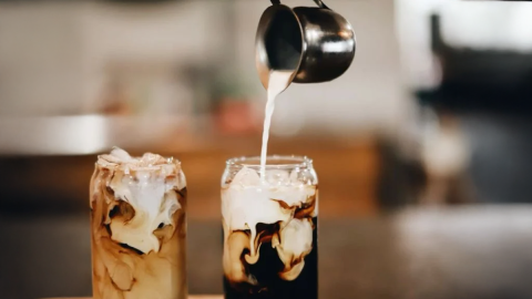 Steamed milk being poured into two glasses around the height of soda-cans - the strong coffee inside is mixing with the milk.