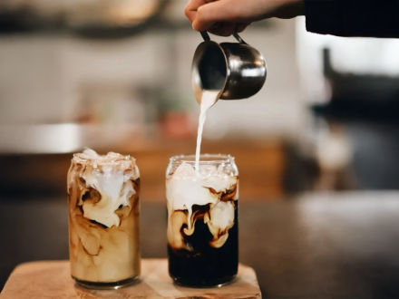 Steamed milk being poured into two glasses around the height of soda-cans - the strong coffee inside is mixing with the milk.