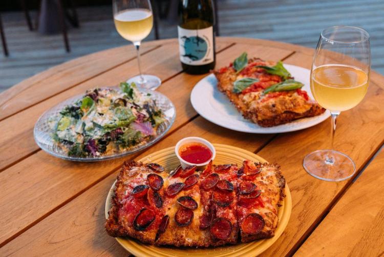 A photo of De La Nonna's pizza and wine, there is a salad on a clear plate and square pizzas with varying toppings.