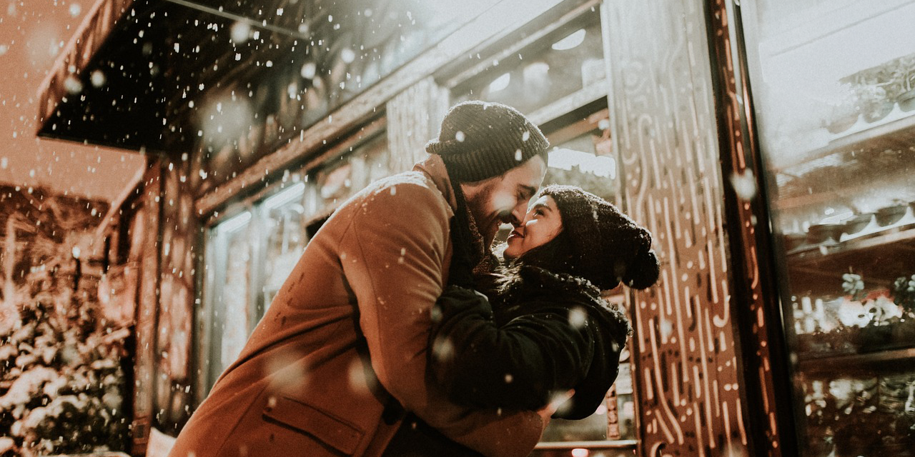 A photo of a couple holding each other lovingly, smiling in front of a store while the snow falls around them.