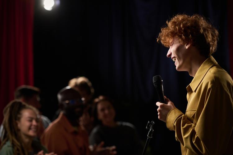 A photo of a male in a yellow shirt holding a microphone, smiling brightly as he speaks in front of a crowd.