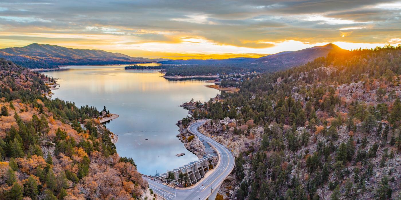A drone shot of Big Bear Lake in fall, the sunset is peaking from behind the mountains and gently reflecting on the lake.