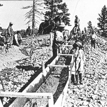 A black and white photo of prospectors panning for gold using a man made stream.