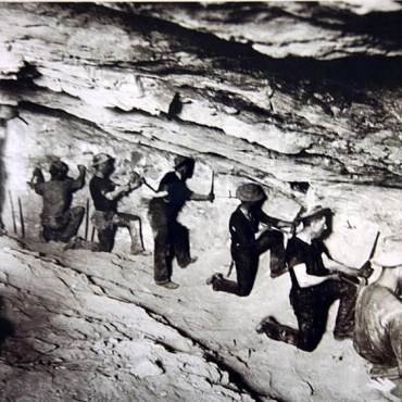A black and white photo of a group of prospectors mining for gold in a cave, they using chisels to search the rock.