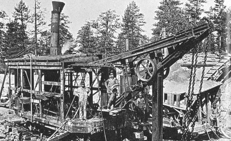 A black and white photo of workers on a machine with chains and belts, the exhaust coming out of the top of the machine.