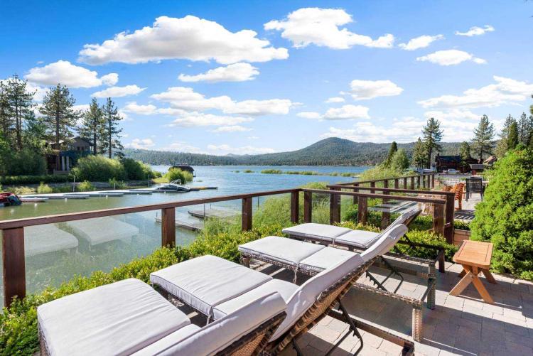 Four chaise lounges sit facing Big Bear Lake from a vacation home deck. Forested mountains and blue sky are shown.