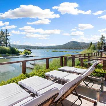 Four chaise lounges sit facing Big Bear Lake from a vacation home deck. Forested mountains and blue sky are shown.