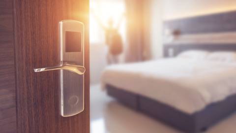 A close up of a hotel room's entrance, out of focus in the background is a bed and a person standing opening the curtains.