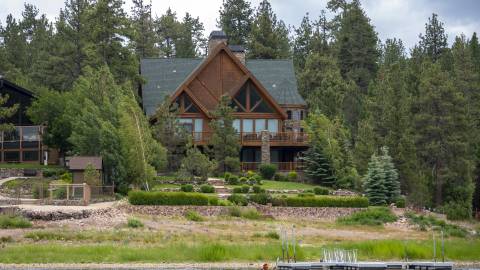 A beautiful brown house sits on the shore of Big Bear Lake. A kayaker paddles by in the water.
