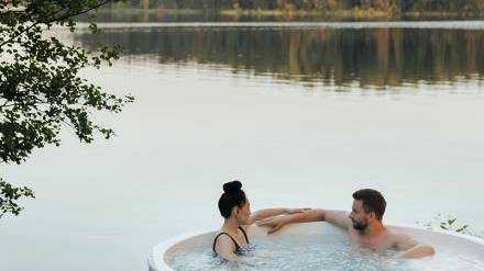 A couple relaxing in a hot tub overlooking the lake, the trees in the distance gently reflecting on the lake.