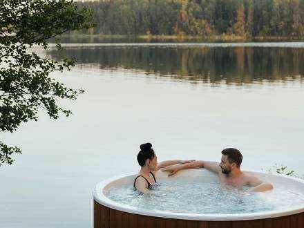 A couple relaxing in a hot tub overlooking the lake, the trees in the distance gently reflecting on the lake.