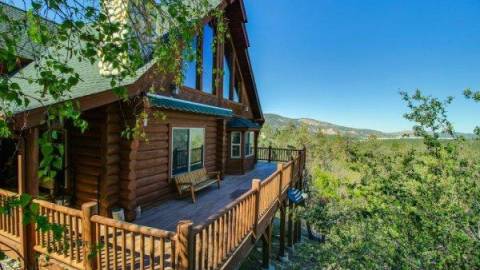 A log cabin surrounded by green oak trees. A large deck wraps around the home.