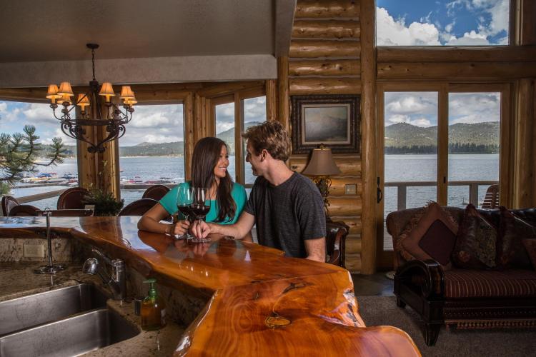 A couple sits at a wooden counter with glasses of wine inside a cabin with a view of Big Bear Lake outside the windows.
