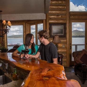 A couple sits at a wooden counter with glasses of wine inside a cabin with a view of Big Bear Lake outside the windows.