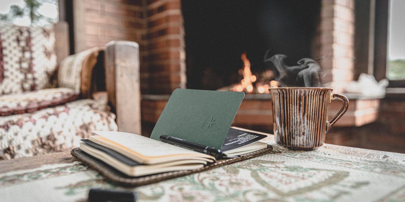 A close up photo of a note book and a steaming hot drink on a table, in the background the fireplace has a lit fire.